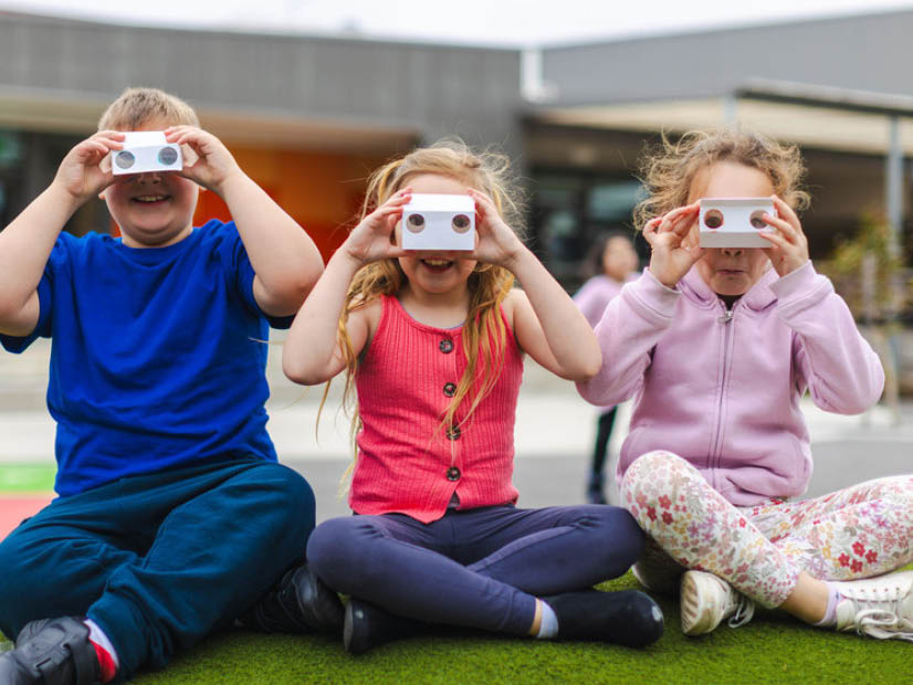 Three kids sitting down outside at Barton OSHC holding crafted viewfinders to their eyes