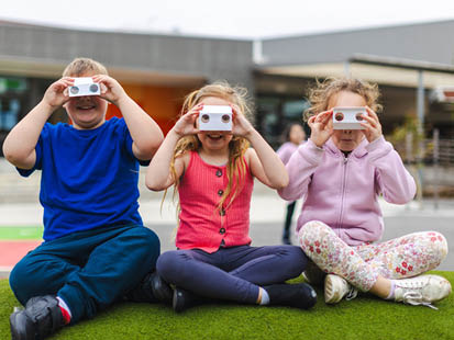 Three kids sitting at outside school hours care holding up crafted viewfinders