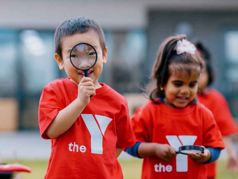 Boy outside holding magnifying glass up to his eyes with girl next to him