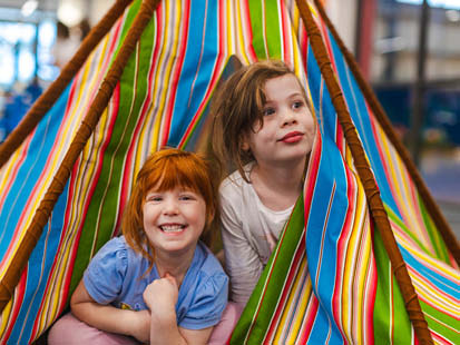 Two girls smiling peeking out the doorway of striped teepee tent inside OSHC room