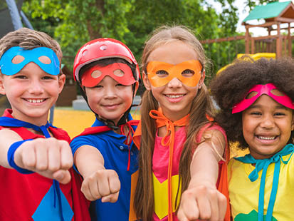 Four kids smiling, dressed in super hero costumes, standing in a row with their arms outstretched like superman