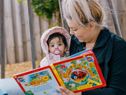 Childcare educator sitting outside holding a baby and reading a book to the child