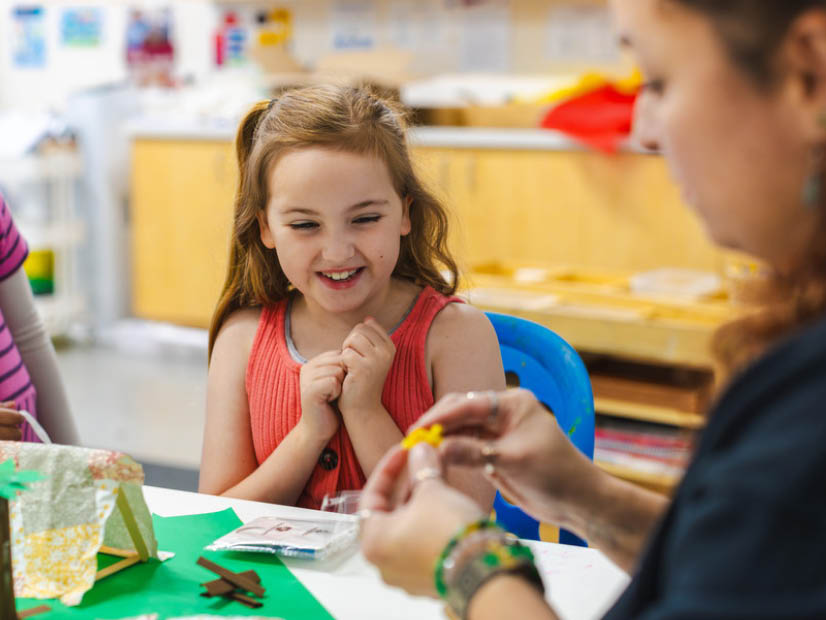 Girl sitting at table delighted watching OSHC educator show her how to craft something