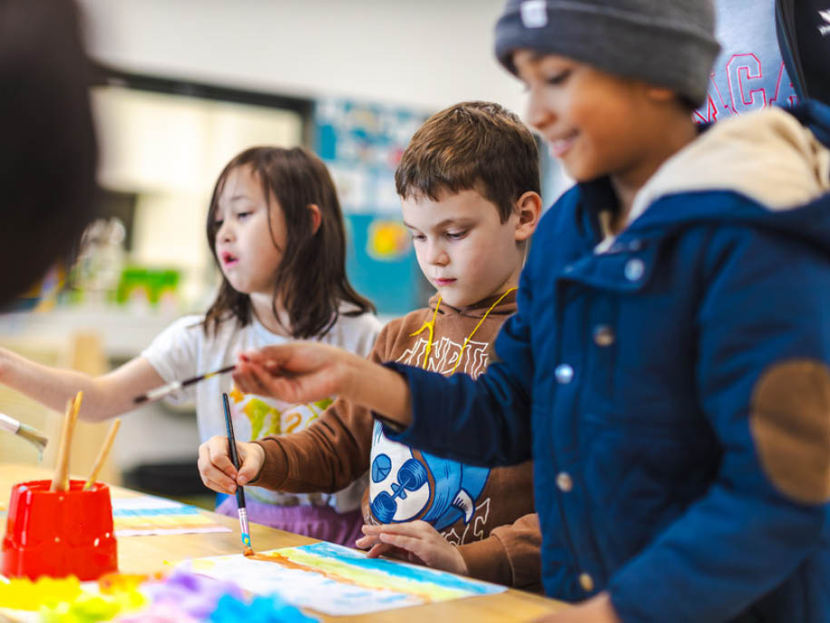 Two boys and a girls at a table painting and OSHC in background watching