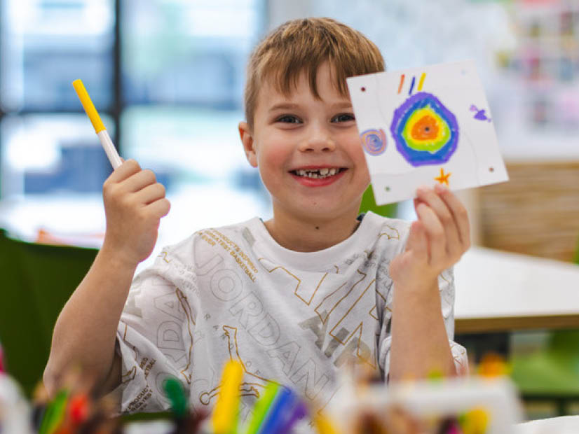 Boy smiling sitting at a table holding up his drawing at holiday program